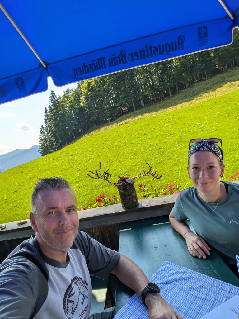 Selfie von Robert und Eva auf der Terrasse einer Alm in den Chiemgauer Alpen, dahinter eine grüne Bergwiese