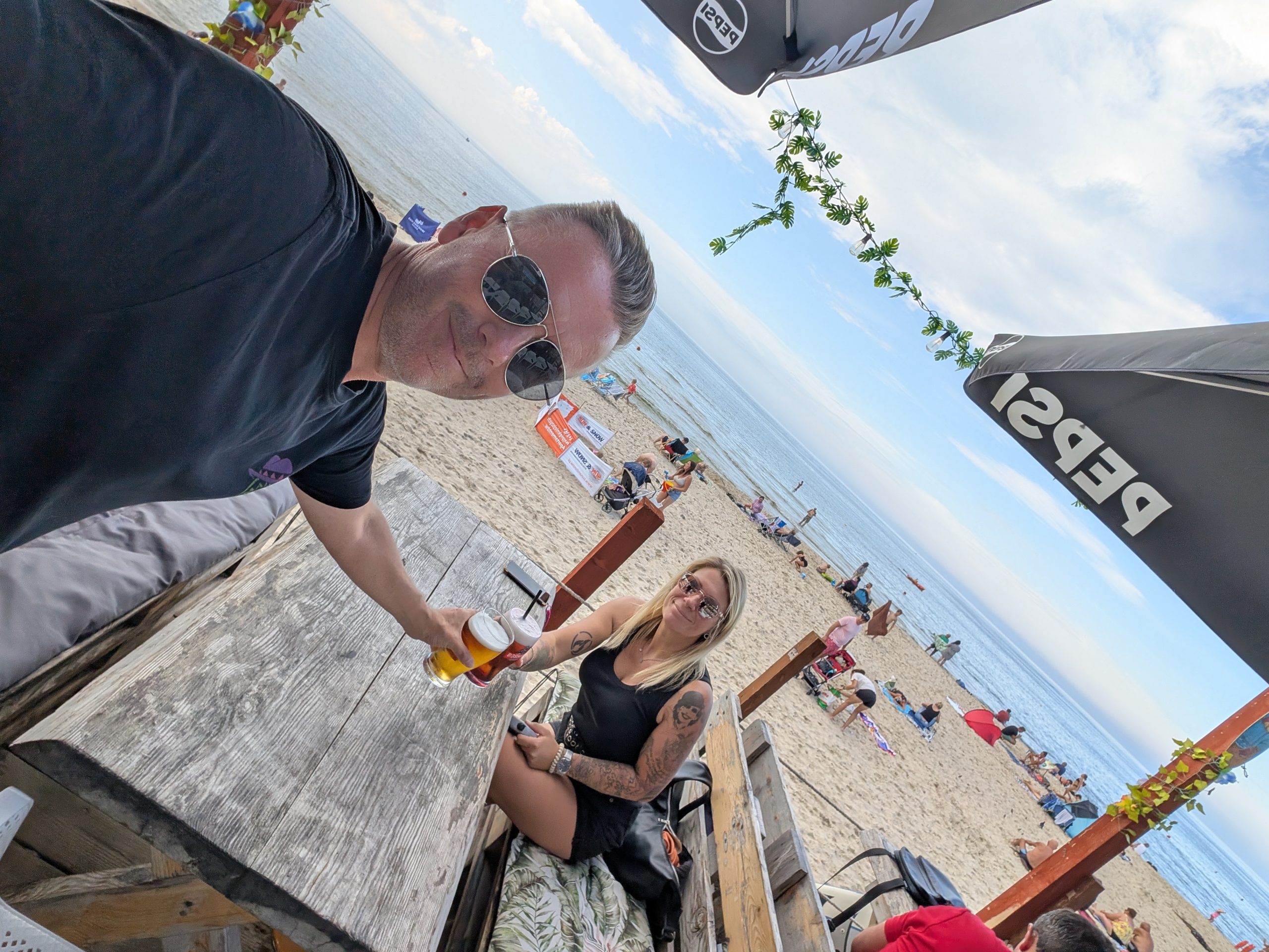 Robert schenkt an einem Holztisch am Strand von Misdroy ein Bier ein, im Hintergrund Sonnenschirme und Ostsee