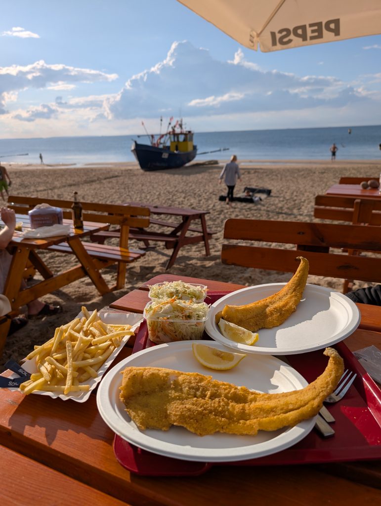 Zwei Teller mit gebackenem Fisch und Pommes auf einem Tablett, dahinter ein Fischkutter am Strand von Misdroy