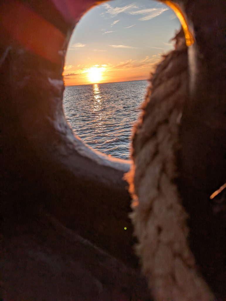 Blick durch eine runde Öffnung in der hölzernen Bordwand auf die untergehende Sonne über der Ostsee