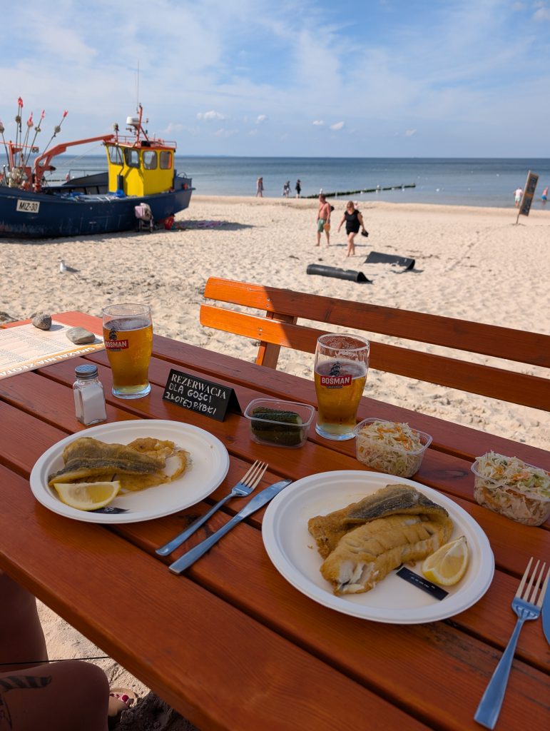 Zwei Teller Fisch und zwei Gläser Bier auf einem Holztisch, dahinter Kutter und die Ostsee