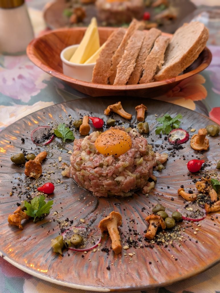 Beef Tatar auf einem Keramikteller, angerichtet mit rohem Eigelb, Pfifferlingen, Kapern, Radieschen und Kräutern, dahinter geröstetes Brot in einer Holzschale