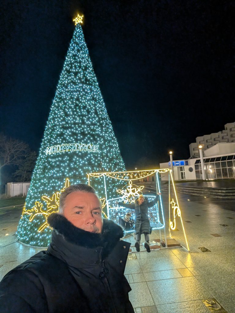 Robert macht ein Selfie vor dem beleuchteten Weihnachtsbaum mit Międzyzdroje-Schriftzug, dahinter posiert Eva in einem Lichterrahmen
