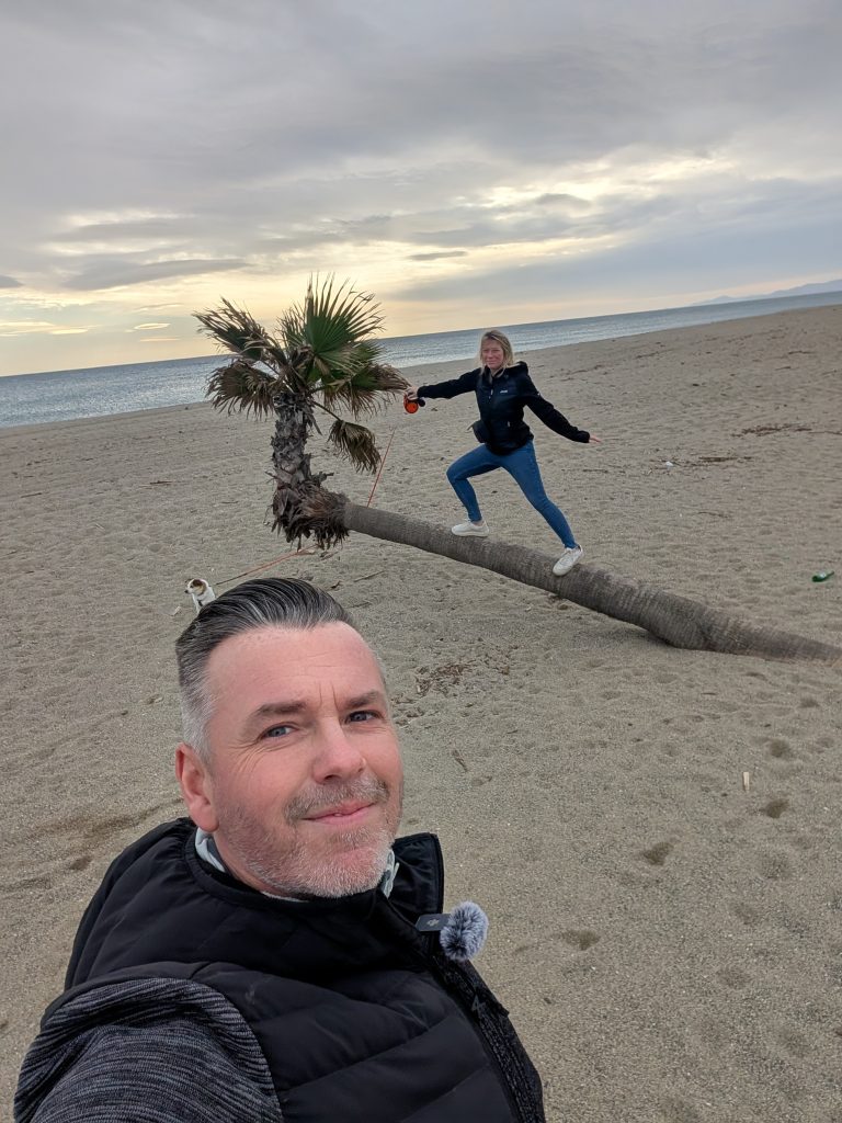 Robert beim Selfie am Strand von Le Barcarès, dahinter balanciert Eva auf einer vom Wind schief gewachsenen Palme