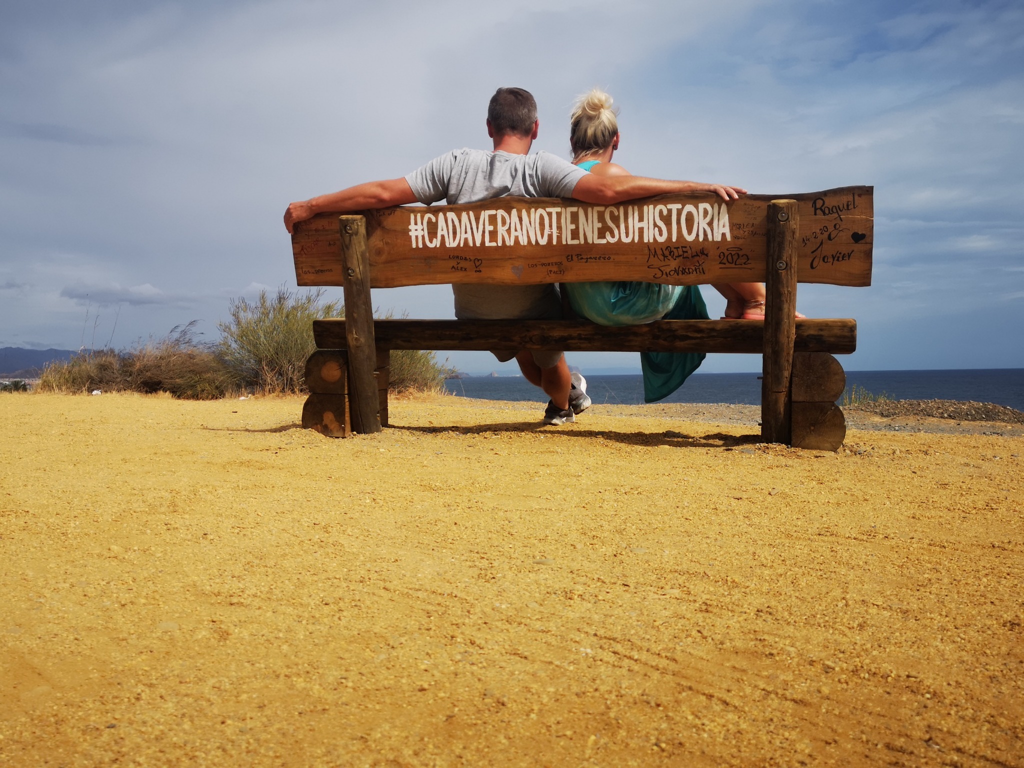 Eva und Robert von hinten auf einer Holzbank mit dem Schriftzug „#CadaVeranoTieneSuHistoria“, Blick aufs Meer