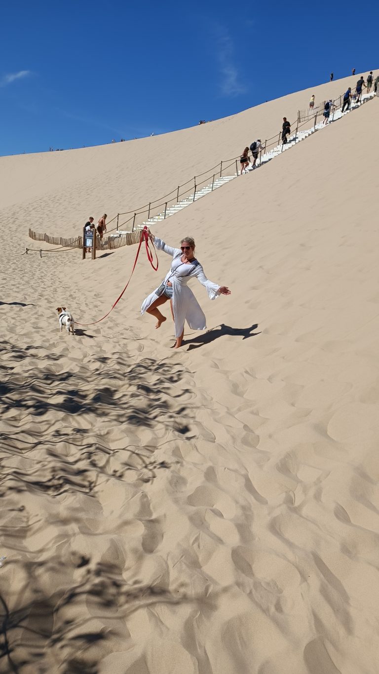 Ausgelassener Sprung am Fuß der Dune du Pilat mit Hund an der Leine, im Hintergrund die Holztreppe