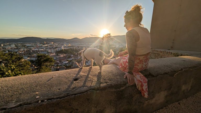 Eva sitzt mit Hund Tyson auf der Mauer am Grazer Schlossberg, die Sonne geht über der Stadt unter