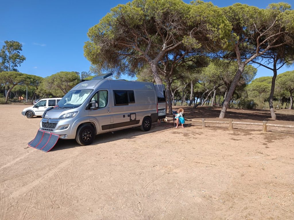Kastenwagen mit Solarmatte im Schatten der Pinien in Isla Cristina, Eva sitzt auf der Holzbank