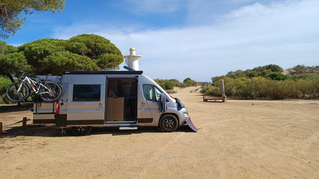 Unser Kastenwagen mit Fahrrädern unter Pinien in Isla Cristina, dahinter ein weißer Turm, September 2022