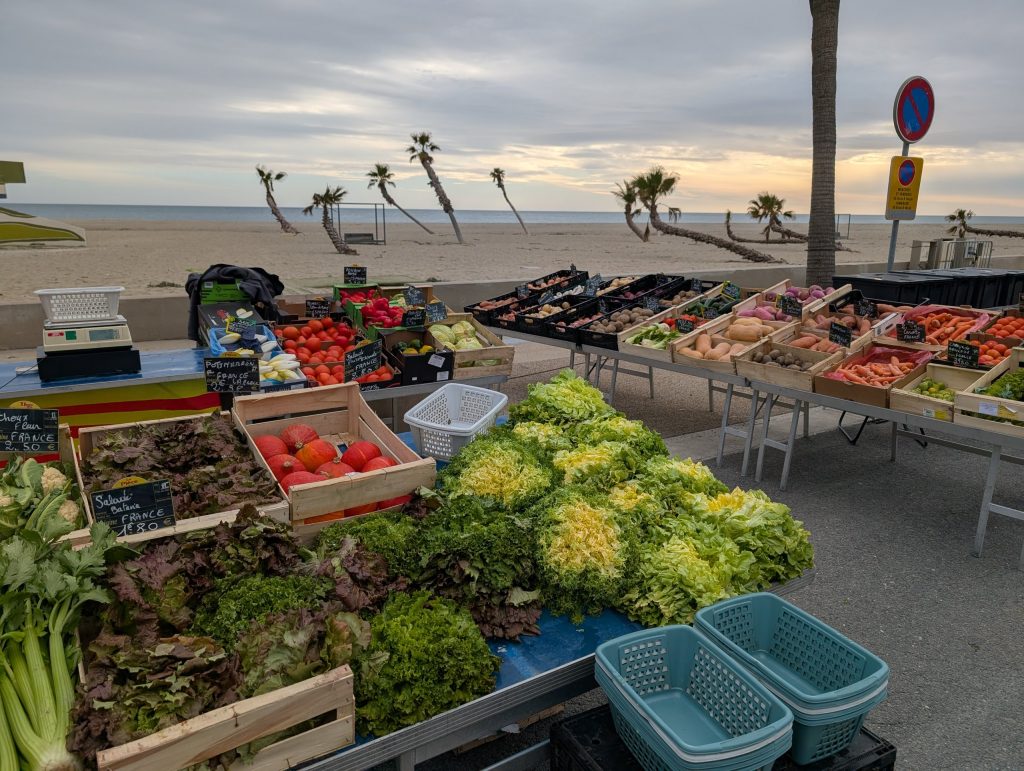 Marktstand mit Salat, Tomaten und Gemüse an der Promenade, dahinter schiefe Palmen am Strand
