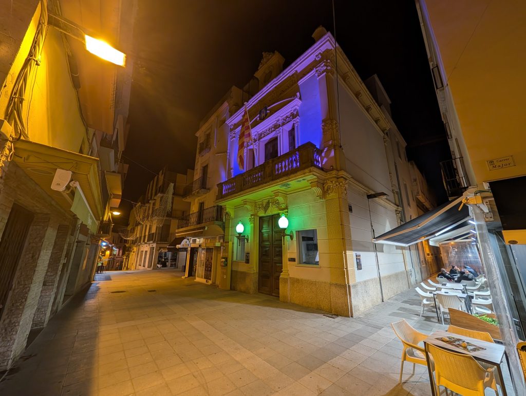 Beleuchtetes altes Stadthaus mit Balkon in der Innenstadt von Palamós am Abend, davor Tische einer Bar