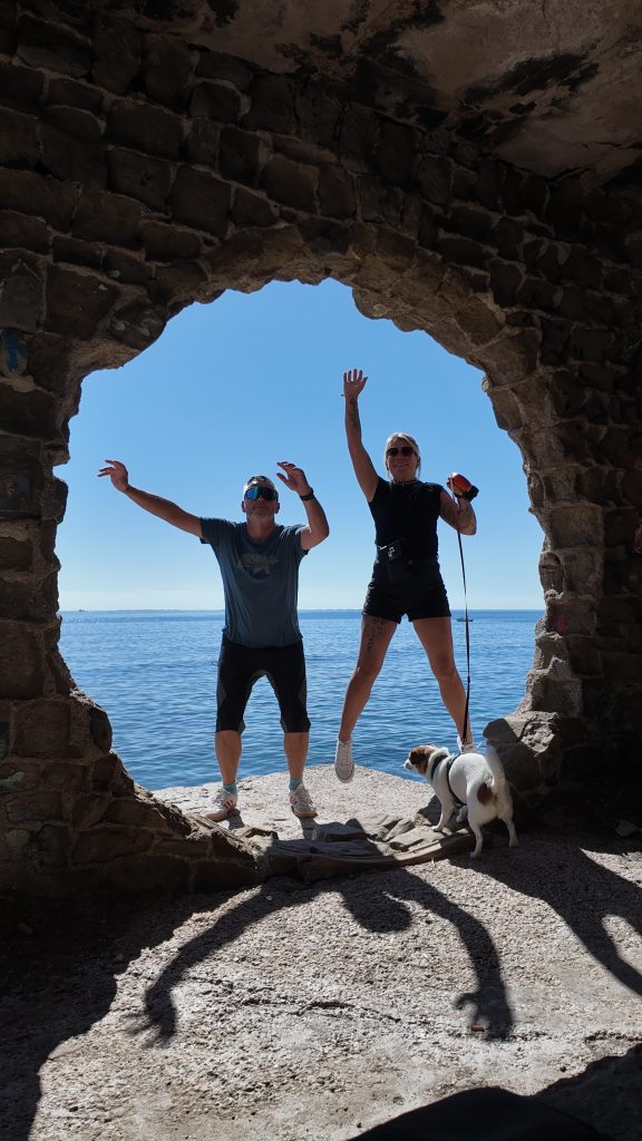 Robert und Eva stehen jubelnd mit erhobenen Armen in einem Fensterbogen des Social Club bei Calp, Tyson davor, dahinter das Meer