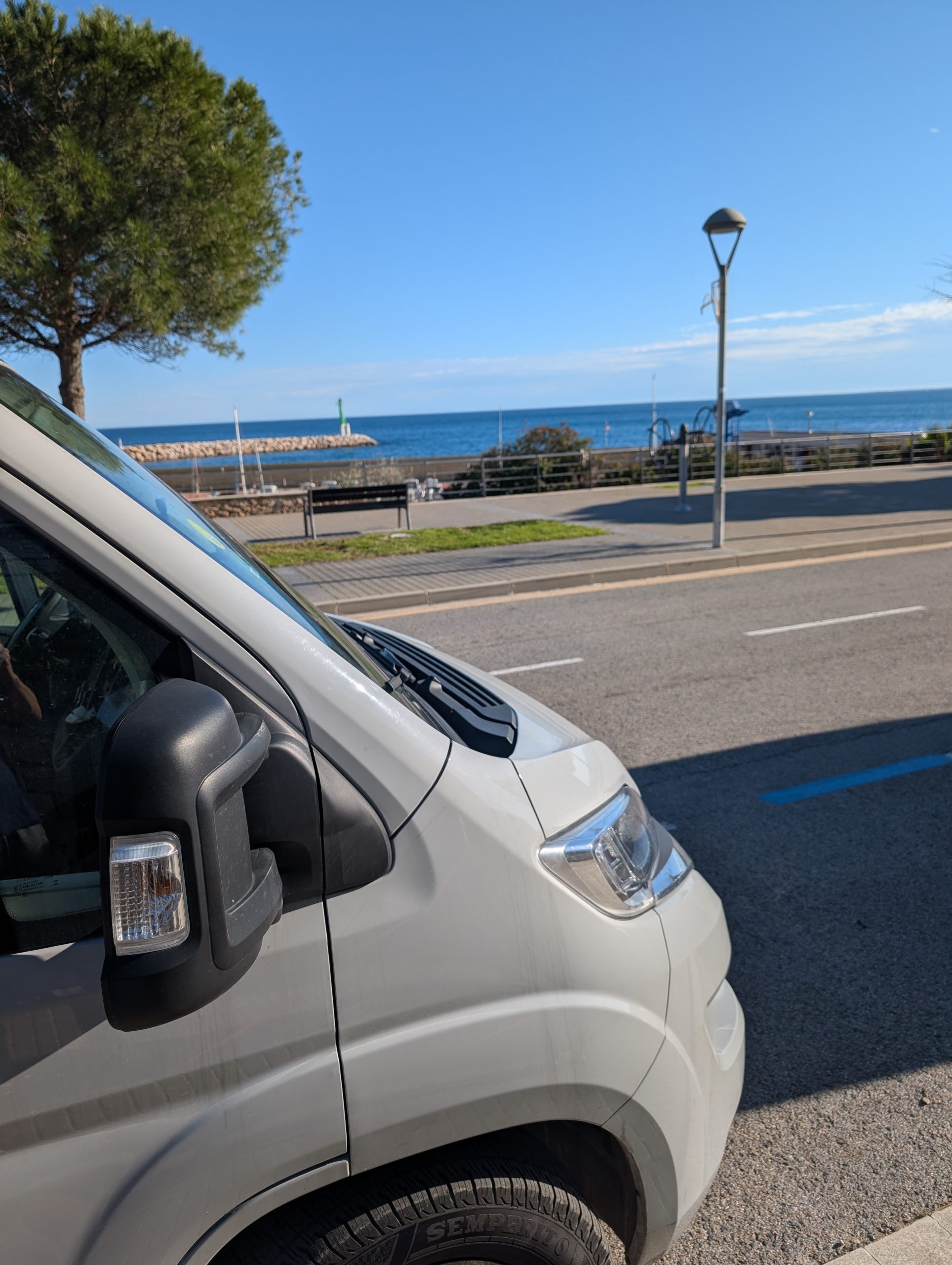 Weißer Kastenwagen an einer Straße in L'Ametlla de Mar, dahinter Promenade und Meer