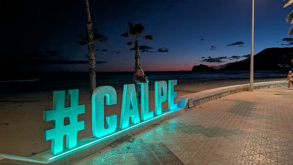 Eva am beleuchteten Schriftzug #CALPE an der Strandpromenade von Calp, kurz nach Sonnenuntergang, dahinter Palmen und Meer