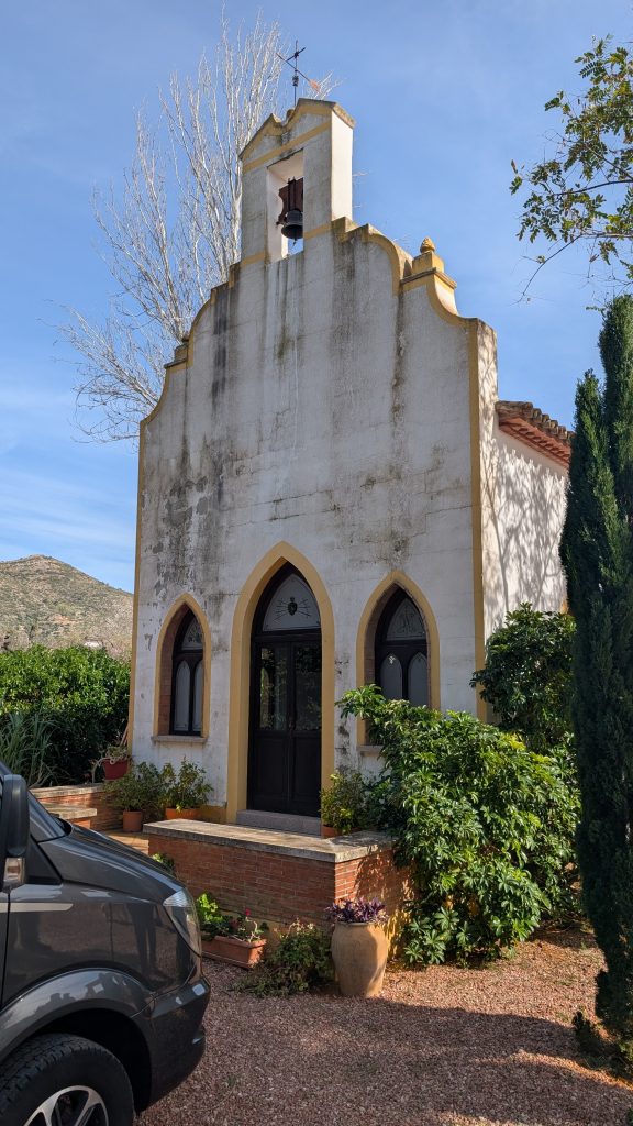 Kleine weiße Kapelle mit gelb abgesetzten Rändern und Glockengiebel auf dem Stellplatz in Alcalalí