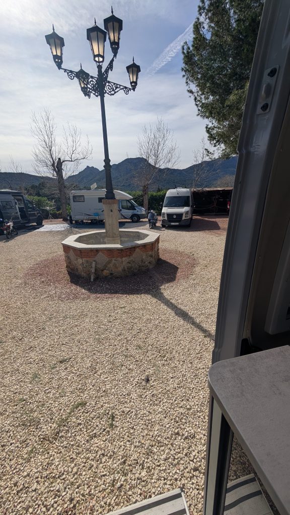 Kiesplatz mit Brunnen und alter Laterne auf dem Stellplatz in Alcalalí, dahinter Wohnmobile und Berge